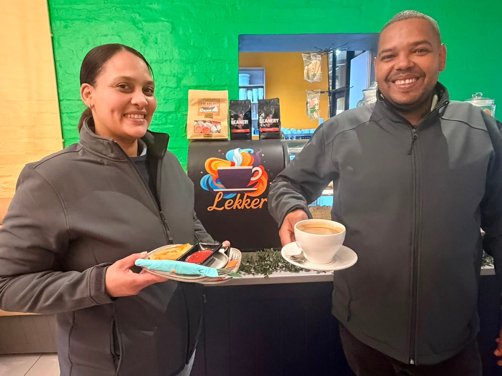 The Lekker Koffiewinkel team behind the counter, holding a plate of food and a cappuccino in front of the Lekker logo